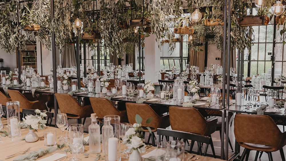 Eleganter, festlich dekorierter Raum mit langen Tafeln, braunen Stühlen, weißen Kerzen, Gläsern und hängendem Grün.