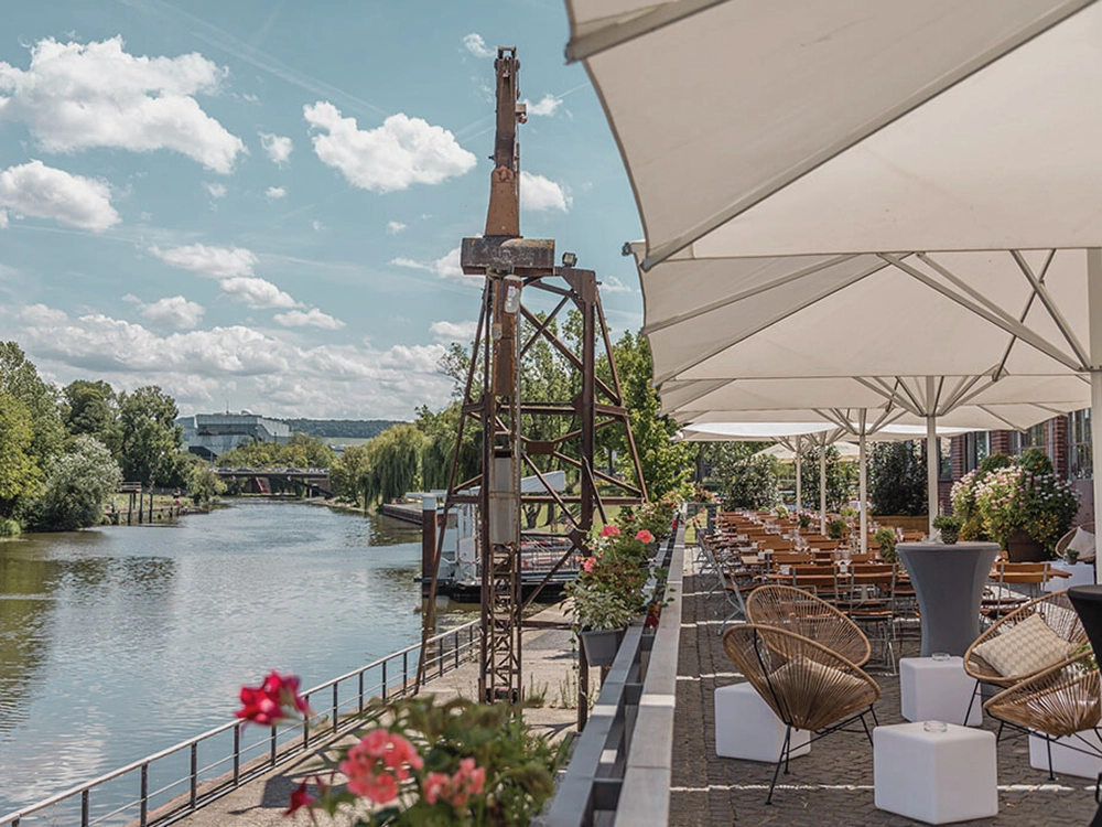 Terrasse mit Tischen und Stühlen am Flussufer unter Sonnenschirmen, umgeben von Pflanzen, ruhige Atmosphäre.