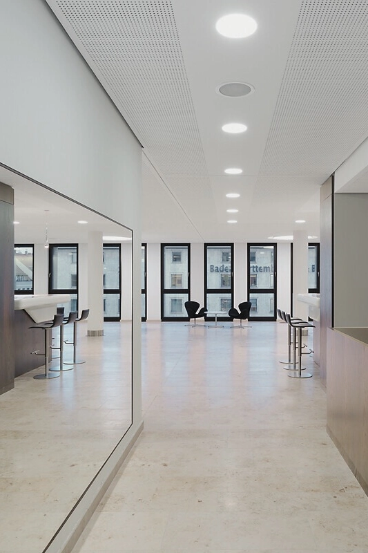 Bankhaus_Eingang Moderner, leerer Büroraum mit großen Fenstern, weißen Wänden und Hochstühlen an Stehtischen, im Hintergrund schwarze Sesseln.