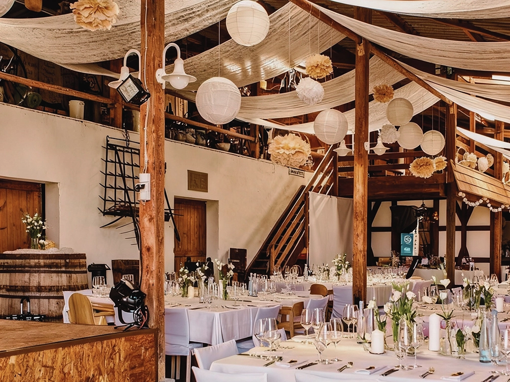 Rustikaler Hochzeitsempfang in einer Scheune mit gedeckten Tischen, Blumenarrangements und hängenden Papierlaternen.