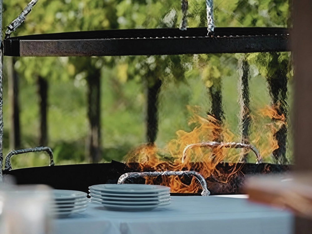 Ein Grill feuert im Freien, davor steht ein Stapel weißer Teller auf einem Tisch, im Hintergrund grüne Bäume.