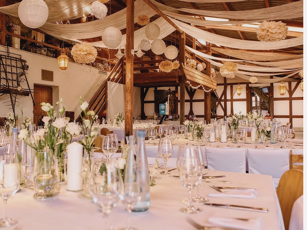 Rustikale Halle dekoriert für Hochzeit mit weißen Tischdecken, Blumenarrangements und hängendem Papierlampions.
