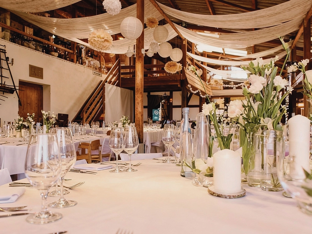 Eleganter Hochzeitssaal mit festlich gedeckten Tischen, weißen Tischdecken, Blumenarrangements, Kerzen und hängenden Papierlaternen.