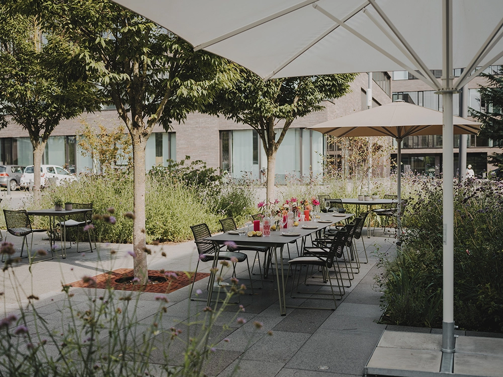 Gedeckter Terrassentisch mit Blumen und Getränken unter Sonnenschirmen, umgeben von Bäumen und Pflanzen in städtischem Umfeld.