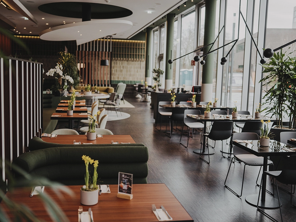 Modernes Restaurant mit grünen Sofas und Tischen, dekoriert mit Pflanzen, großen Fenstern rechts, helle und einladende Atmosphäre.
