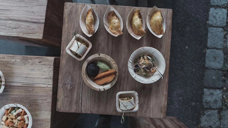 Holztisch mit verschiedenen Speisen in Schalen, darunter Teigtaschen, vegetarische Häppchen und Salat. Draufsicht.