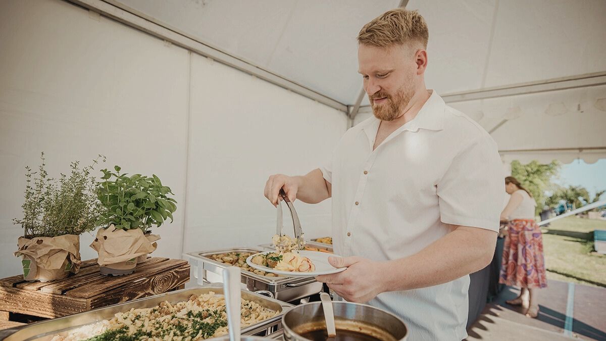 VOLTINO_Cateringstation Mann schöpft Essen an Buffet unter Zelt, mit Kräutern auf Holzregal; draußen sonnig, entspanntes Ambiente.