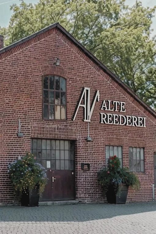 Ziegelgebäude mit Schriftzug Alte Reederei, Holztür, Fenstern, zwei Pflanzenkübeln, Bäume im Hintergrund, bei Tageslicht.