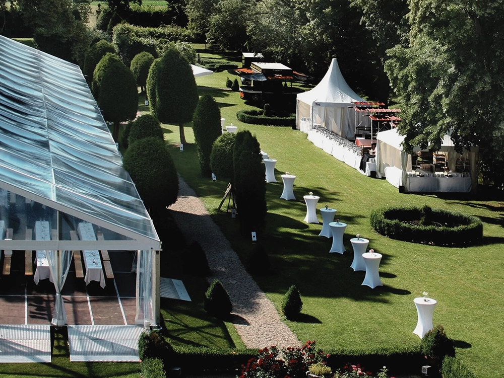 Grüner Garten mit Festzelt und Stehtischen, gesäumt von Bäumen, vorbereitet für eine Veranstaltung bei sonnigem Wetter.