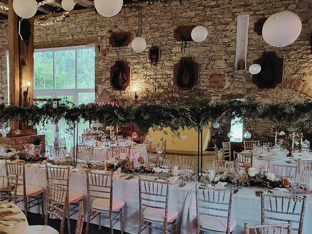 Festlich dekorierter Hochzeitsempfang in rustikalem Saal mit langen, blumengeschmückten Tafeln, Holzstühlen und Steinwänden.