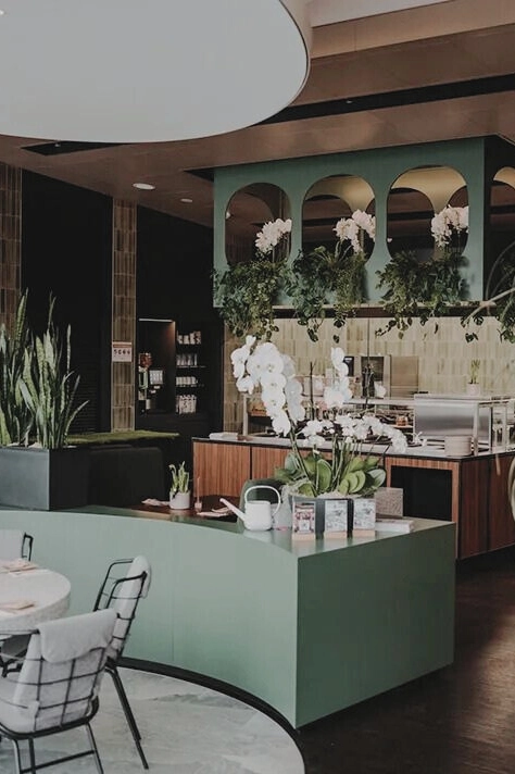 Modernes Café-Interieur mit grüner Theke, Orchideen und hängenden Pflanzen; gemütliche Atmosphäre mit eleganten Möbeln.