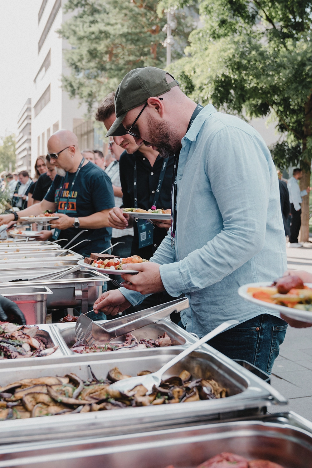 Männer stehen in einer Reihe bei einem Buffet im Freien, servieren sich Essen auf Tellern, umgeben von Bäumen und Gebäuden.