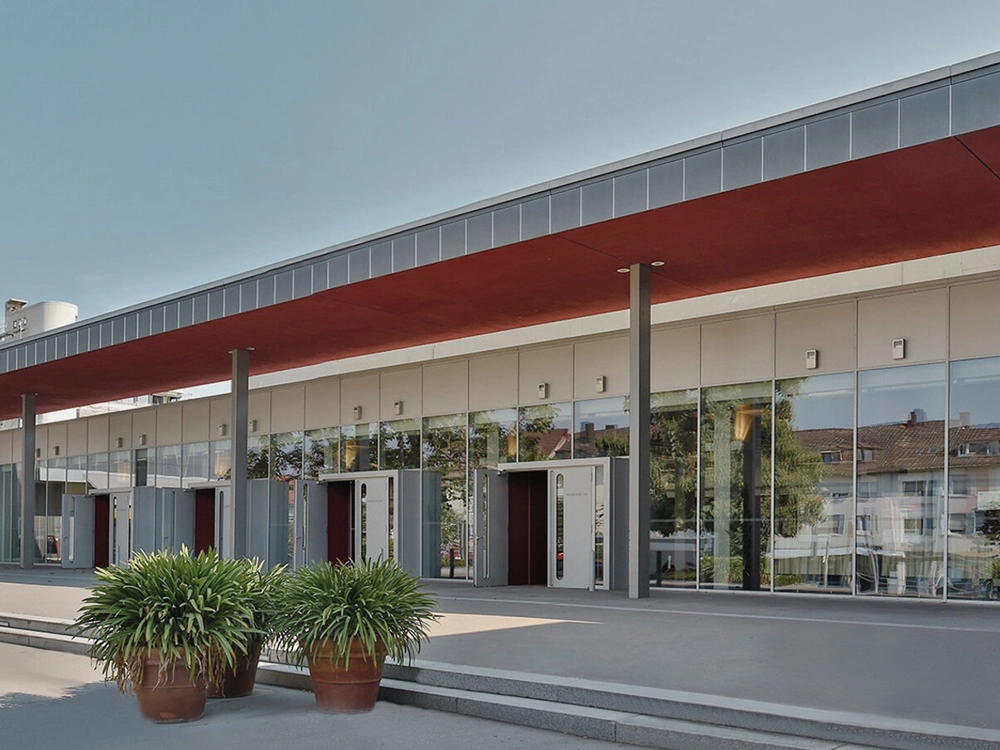 Modernes Gebäude mit Glasfassade und rotem Vordach, zwei große Pflanzen in Töpfen vorne, klarer Himmel im Hintergrund.