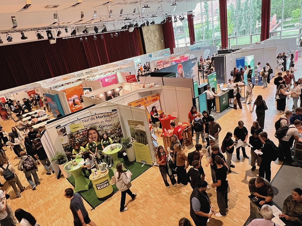 Menschen erkunden eine Messehalle mit verschiedenen Ständen und Roll-Up-Bannern, angeregte Gespräche finden statt.