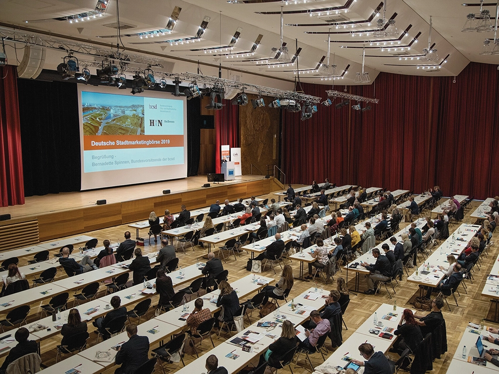 Eine Konferenzhalle mit vielen Teilnehmern, die an Tischen sitzen, und einer großen Leinwand für Präsentationen.