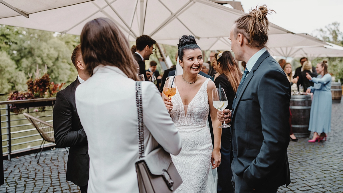 Hochzeitspaar und Gäste im Freien, halten Getränke in der Hand, unter Sonnenschirmen, lächelnd und im Gespräch vertieft.