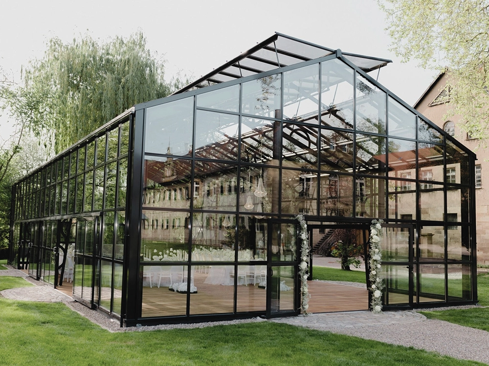Moderne Glasstruktur im Garten, mit gedeckten Tischen und Blumen dekoriert, umgeben von grünen Bäumen und altem Gebäude im Hintergrund.