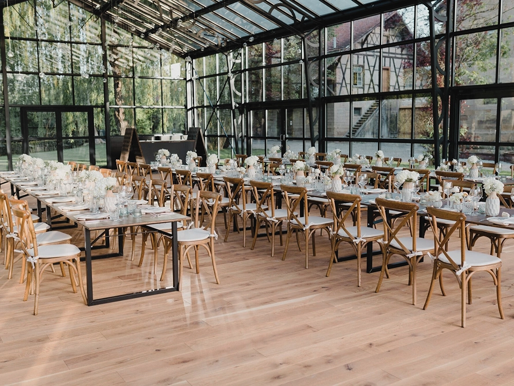 Festlich gedeckte Tische in einem gläsernen Raum mit Holzboden, bereit für ein Event. Florale Dekoration und Holzstühle.