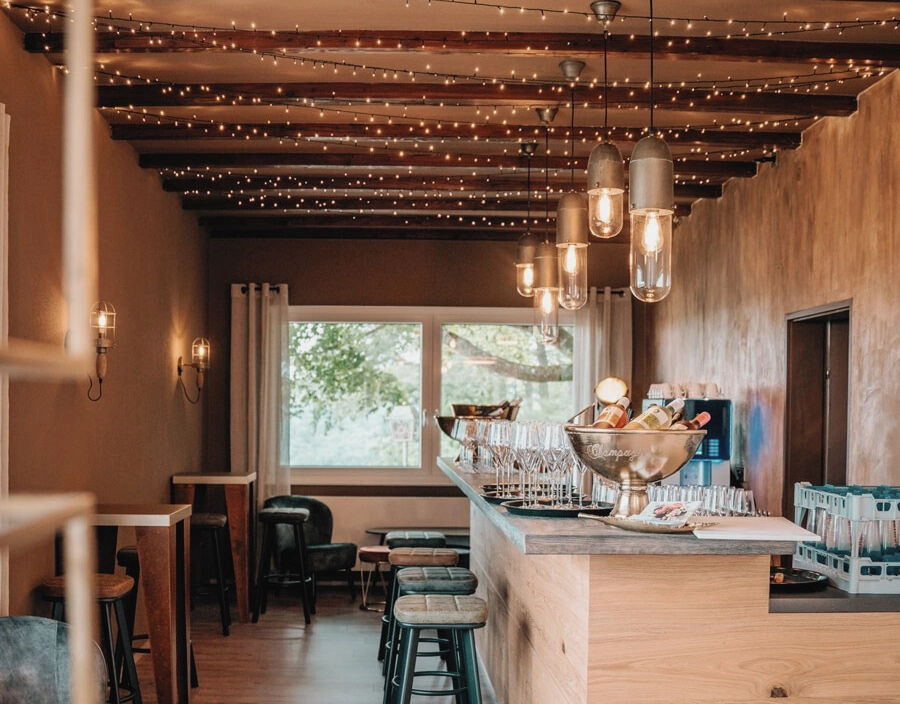 Gemütliche Bar mit Holztheke, Barhockern und Deckenleuchten. Fenster mit Blick ins Grüne, Eiswanne mit Flaschen auf Theke.