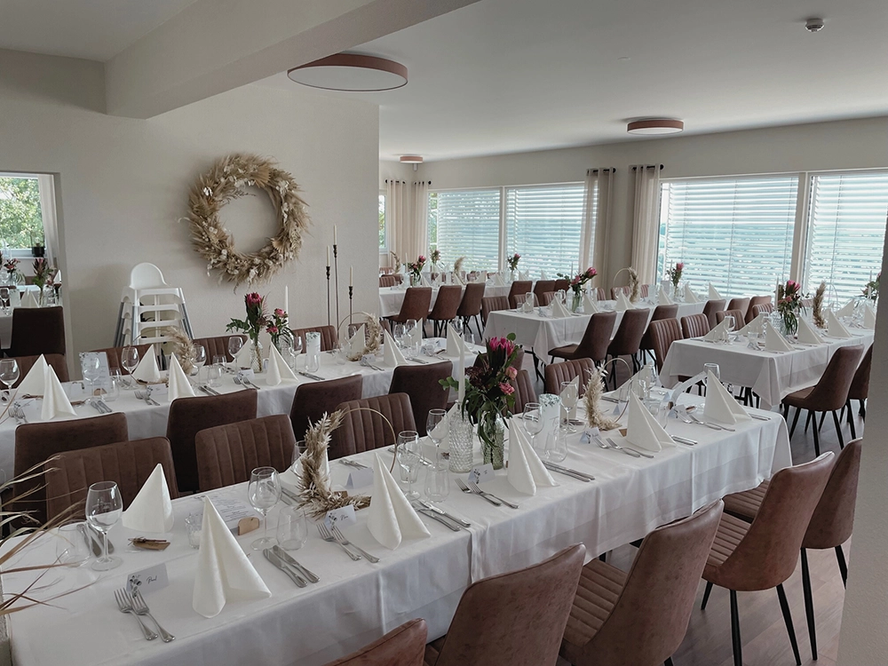 Elegante, weiß gedeckte Tische mit gefalteten Servietten und Blumenarrangements in hellem Raum; Hochstuhl und Wandkranz sichtbar.