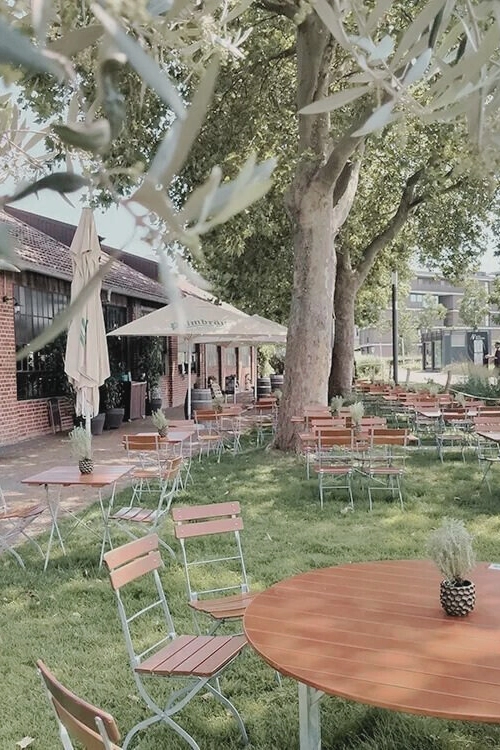 Gartenrestaurant mit leeren Holztischen und Stühlen auf Rasen, großer Baum und Sonnenschirme neben Backsteingebäude.