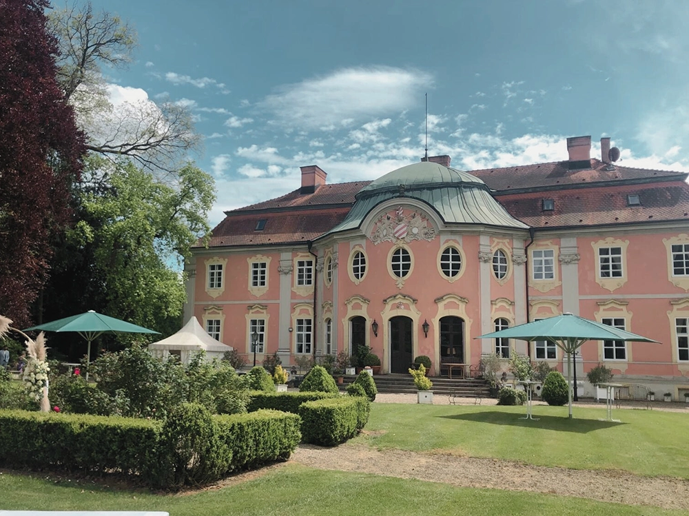 Barockes Gebäude mit rosa Fassade, grüne Gartenanlage und Sonnenschirme im Vordergrund, blauer Himmel im Hintergrund.