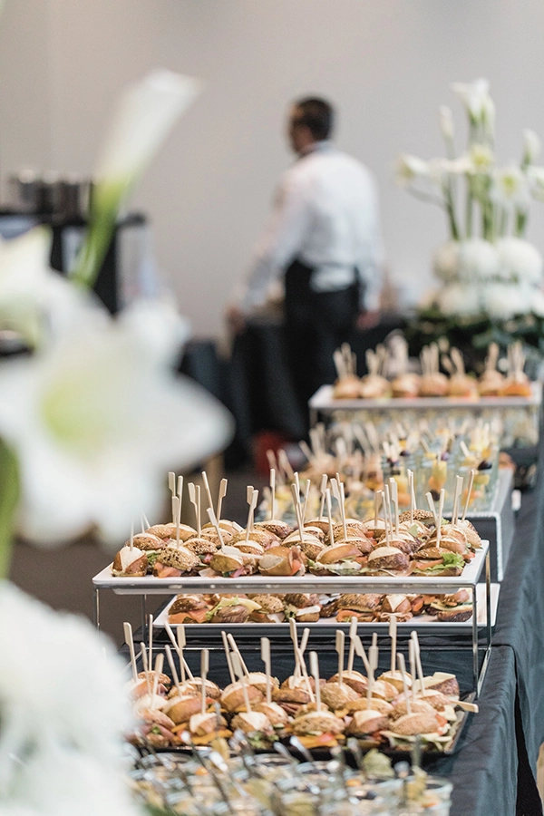 Tablett mit kleinen belegten Brötchen auf einem Buffet; unscharfe Person im Hintergrund, weiße Blumen im Vordergrund.