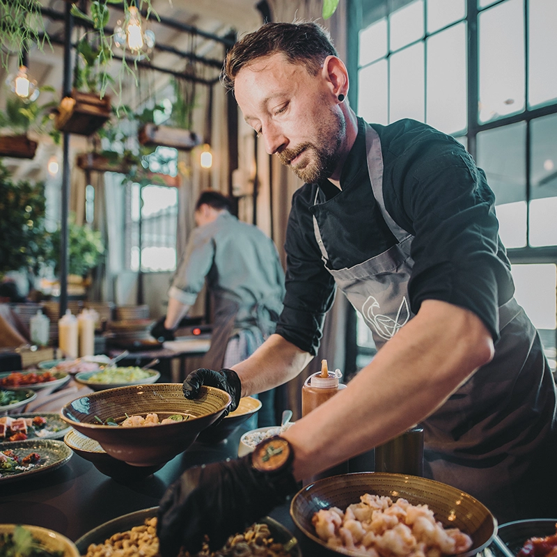 Koch in Restaurant bereitet Speisen an einem Tisch vor. Verwendung von Schüsseln und Zutaten in modernem, lichtdurchflutetem Raum.