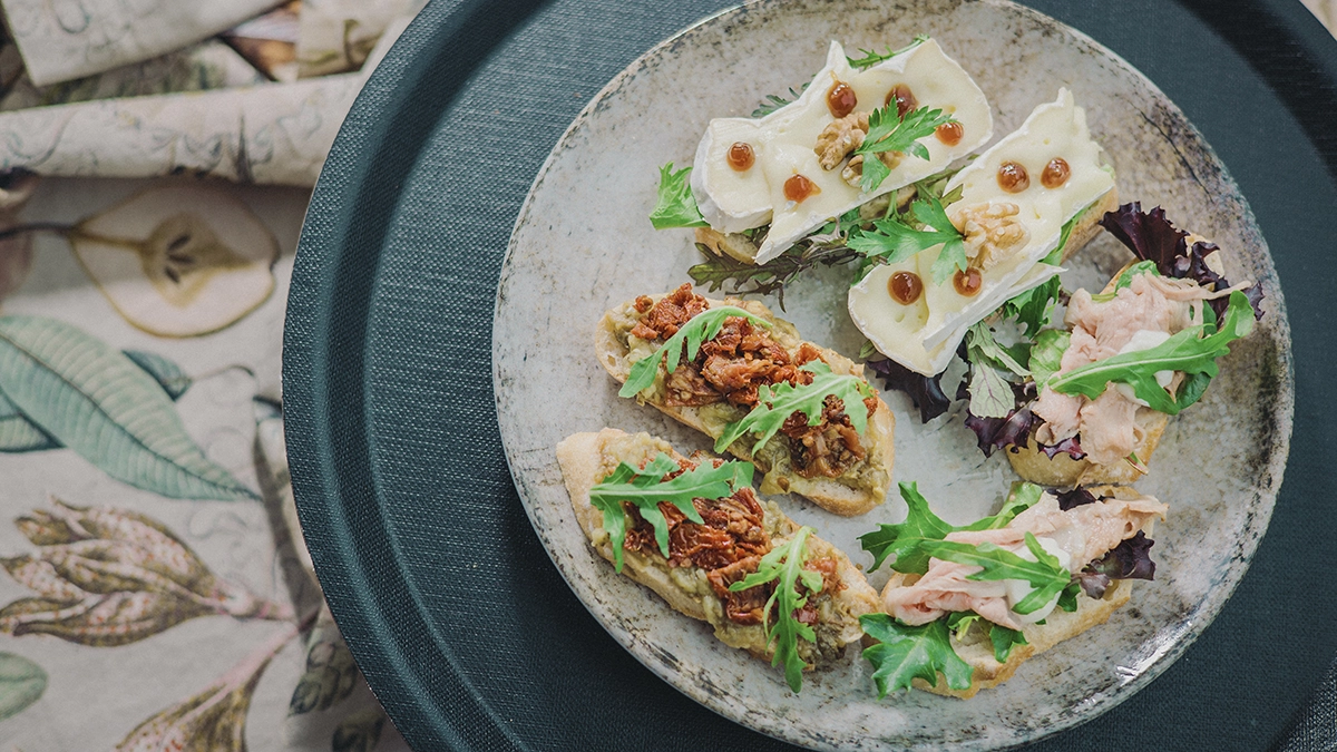 Vorspeisenplatte mit Brötchen: belegt mit Käse, Walnuss, Rucola und Aufstrichen auf einem cremefarbenen Teller.