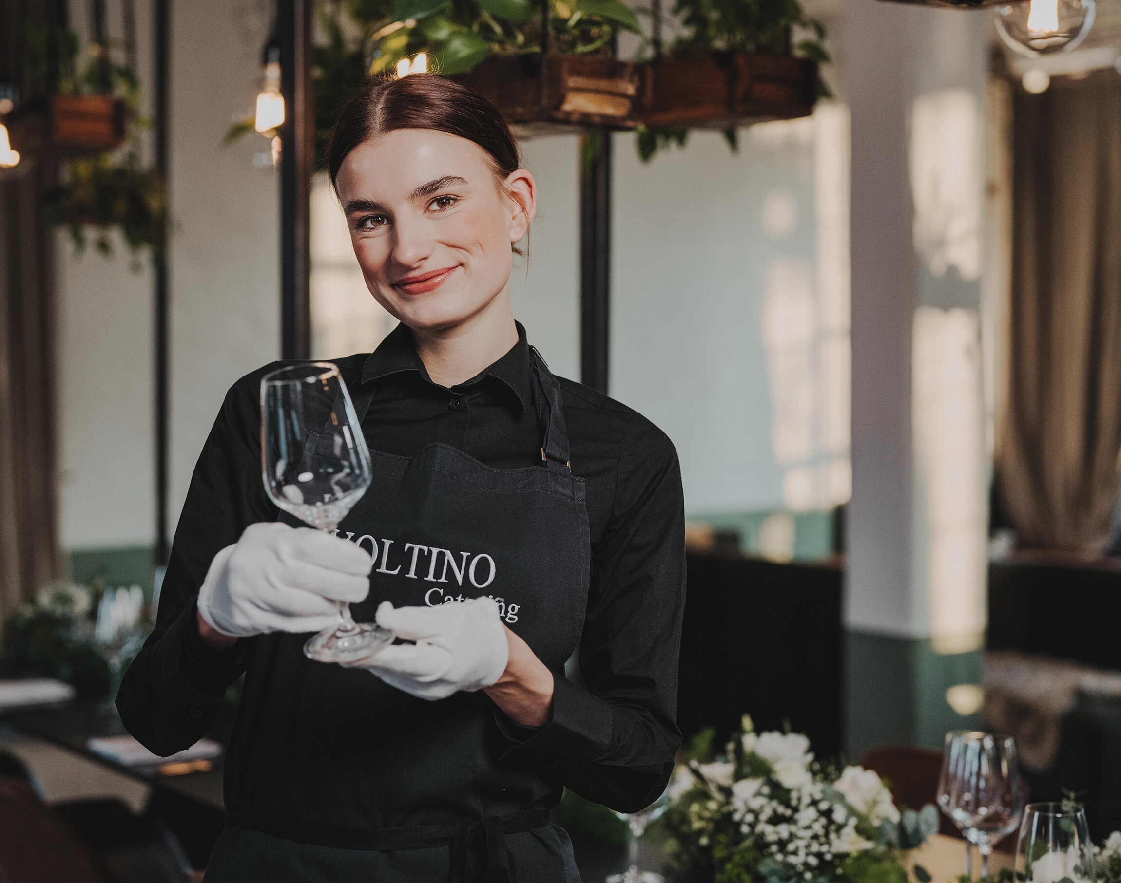Eine lächelnde Bedienung reinigt ein Weinglas im Restaurant, umgeben von Blumen und hängenden Pflanzen.