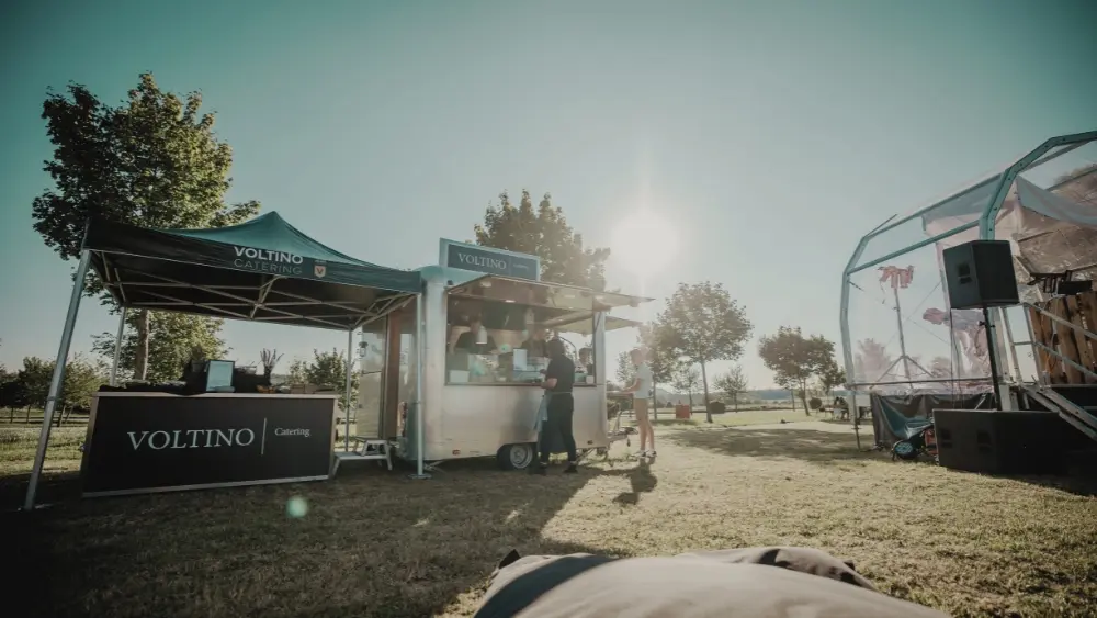 1_3 Foodtruck und Zelt von Voltino Catering auf einer Wiese bei Sonnenschein, umgeben von Bäumen und einem Pavillon im Hintergrund.