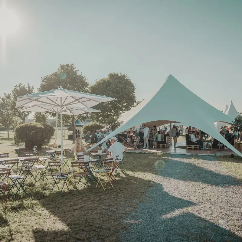 1_3 Outdoor-Event mit Menschen unter einem großen weißen Zelt, leere Tische und Stühle im Vordergrund, sonniger Tag.