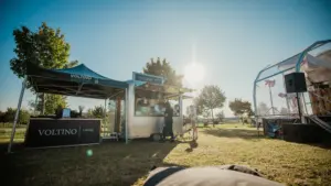 250810_Voltino-272 Essensstand von Voltino Catering auf einer Wiese bei Sonnenschein, Menschen bestellen Essen, Bäume im Hintergrund.