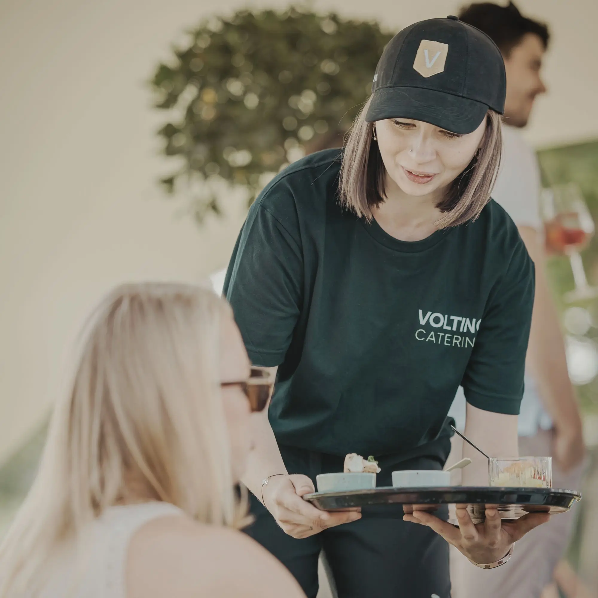 Mitarbeiterin von VOLTINICATERING überreicht Teller mit Speisen an sitzende Person im Freien.