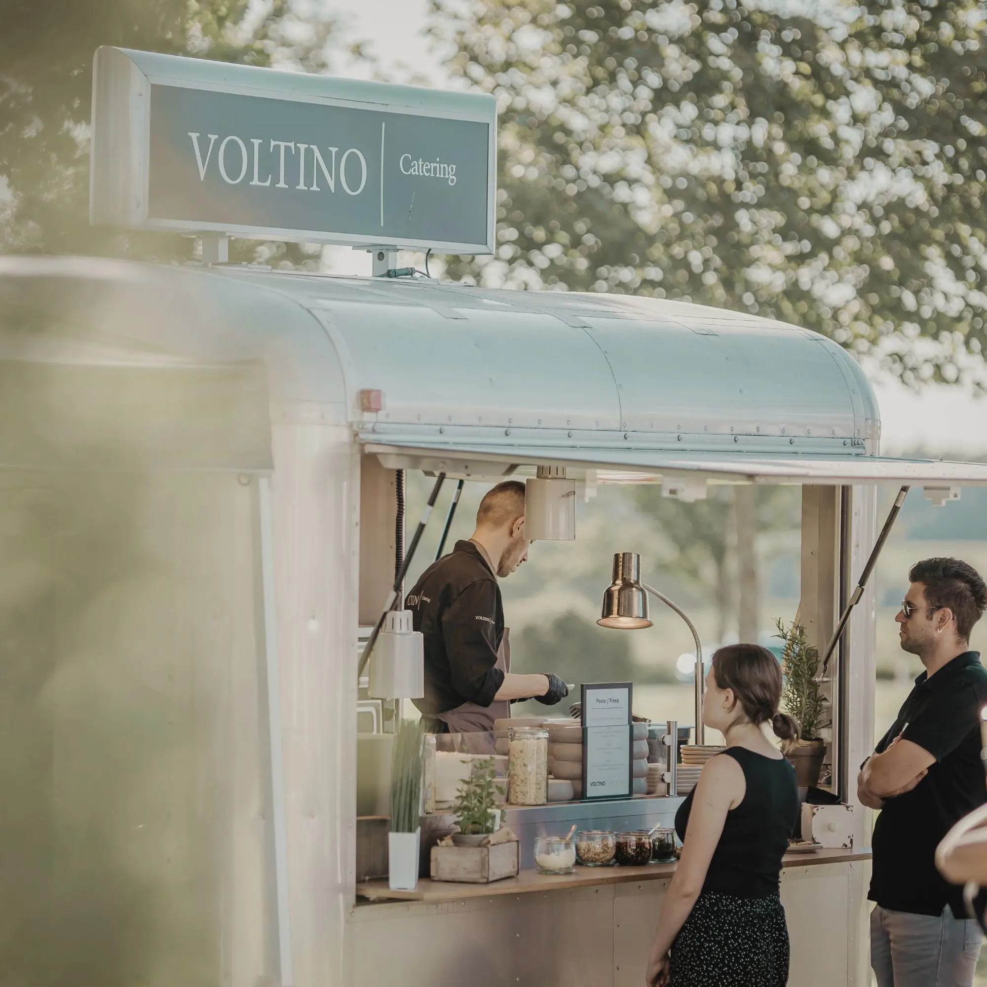Ein Mann bedient in einem Foodtruck zwei wartende Kunden im Freien, unter einem Schild mit der Aufschrift Voltino Catering.