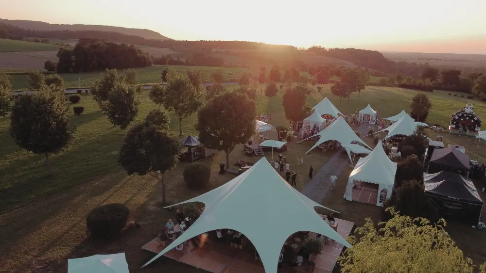 2_3 Freiluftveranstaltung bei Sonnenuntergang mit weißen Zelten auf einer Wiese, umgeben von Bäumen und Menschen in festlicher Stimmung.
