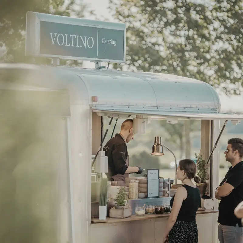 2_3 Ein Mann bedient Kunden an einem Foodtruck namens Voltino Catering in einem sonnigen Park.