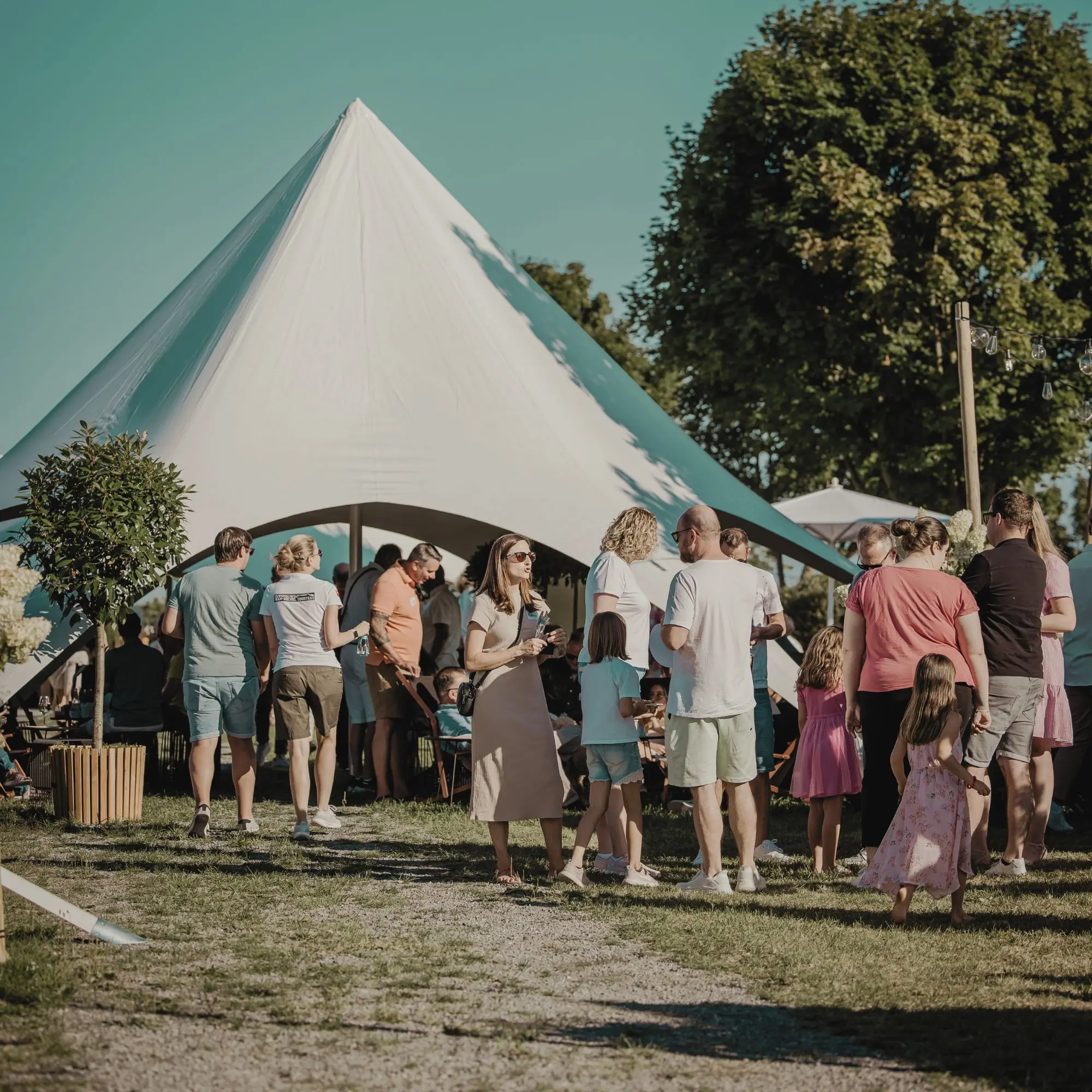 Menschen auf einer Wiese bei einem Fest unter einem großen weißen Zelt im Freien; sommerliche Atmosphäre.