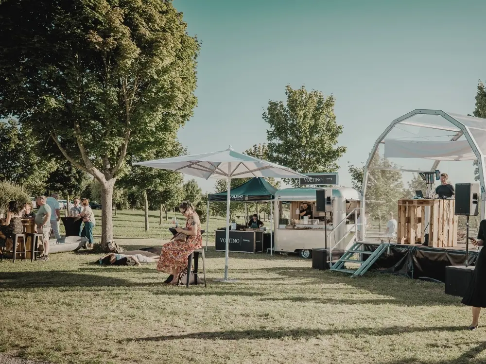 8 Outdoor-Event mit Personen unter Bäumen. Eine Frau liest. Im Hintergrund Bühne, DJ, Verkaufsstand und Besucher.