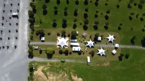 DCIM100MEDIADJI_0117.JPG Luftaufnahme eines Parks mit weißen Zelten auf einer Wiese. Vereinzelte Bäume und parkende Fahrzeuge auf danebenliegendem Asphalt.