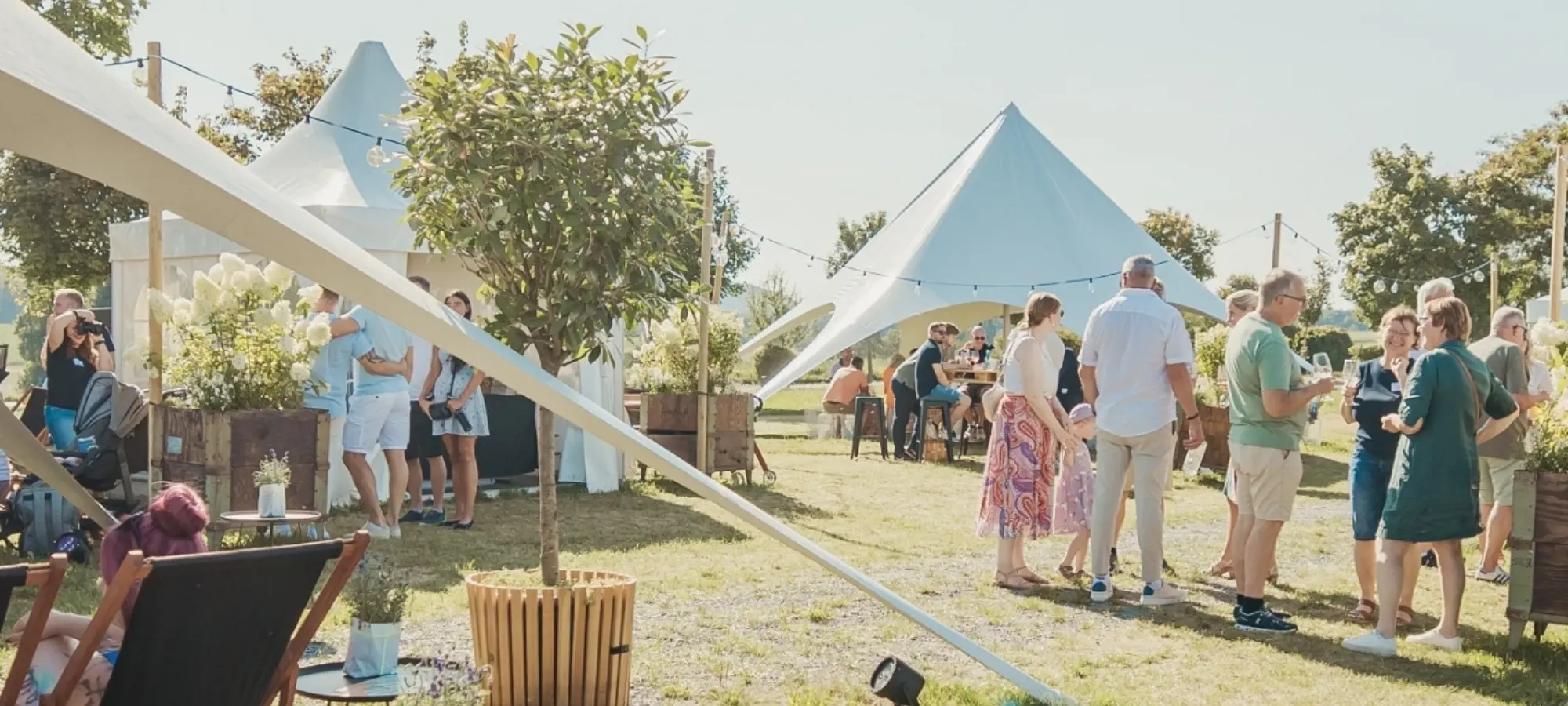 Pop-Up-Fenster_Sommerfeste Menschen in Sommerkleidung versammeln sich bei einem Fest im Freien mit weißen Zelten und Pflanzen, plaudern und genießen die Sonne.